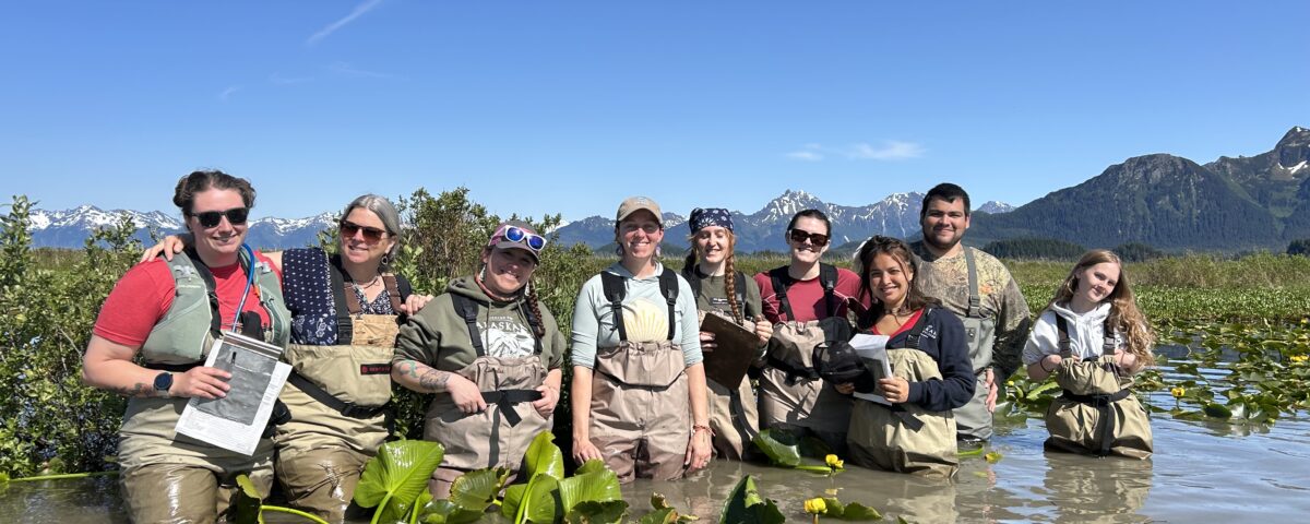 Group standing thigh deep in water full of lily pads. Group is next to a USFS nest island. Group is smiling for a photo. There are 9 people in the photo