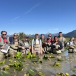 Group standing thigh deep in water full of lily pads. Group is next to a USFS nest island. Group is smiling for a photo. There are 9 people in the photo