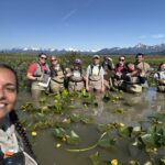 Group standing thigh deep in water full of lily pads. Group is next to a USFS nest island. Group is smiling for a photo. There are 10 people in the photo. One person is taking the photo and is closer to the camera because it is a selfie.