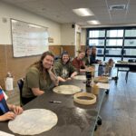 6 peope smiling at the camera. They are sitting at tables inside a classroom. In front of them are supplies to make a drum.