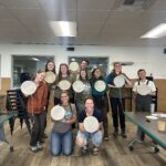 Group of 10 people smiling and looking at the camera. Everyone is holding up an Alaska Native style drum.