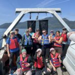 Group of 12 people on a boat in the ocean. Everyone is wearing a lifejacket. Everyone is smiling and looking at the camera.