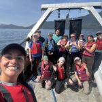 Selfie with a group of 12 people. Everyone is smiling and looking at the camera. Everyone is wearing a life jacket because the photo takes place on a boat