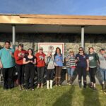 Group of 10 people holding suncatchers, standing in front of a building on a sunny day. The group is smiling for the camerea.