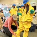 woman helps younger girl put on oil spill response gear (yellow suit and yellow booties with blue hard hat)
