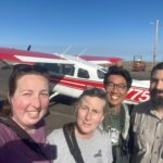 four people (two male and two female adults) smiling for the camera with a red and white plane in the background
