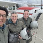 Three adults (two female - right - and one male) in flight suits holding helmets in front of a red and white plane