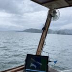 Photo shows the inside of a boat shack. There are glass windows allowing you to see the ocean and mountains in the distance. Inside the shack is a counter where a laptop and notebook are set.