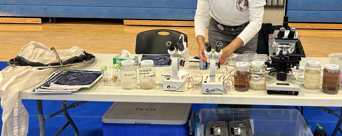 Woman standing at table full of scientific equipment: microscopes, nets, and more.