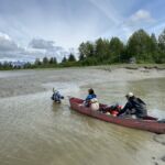 A steward pulls a canoe across a creek, and two stewards ride inside. A steward pulls a canoe across a creek with two stewards riding inside.