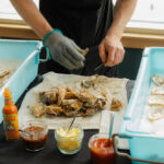 a Man with two blue containers of oysters and various sauces, shucking the oysters with a knife.