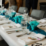 A table with an array of silverware and wine glasses on a white cloth with turquoise decorative paper flowers and a blue table runner.