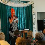 A woman on a stage wearing a squid hat, speaking into a microphone as she reads off a slip of paper.