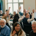 A group of banquet guests raising their hands with white windows in the background.
