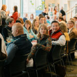 A group of banquet guests seated in black chairs turning to face a stage.