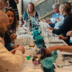 a group of people at a white banquet table with wine glasses and blue flower decorations socializing.