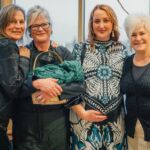 Four smiling women in front of a window in dark clothing.